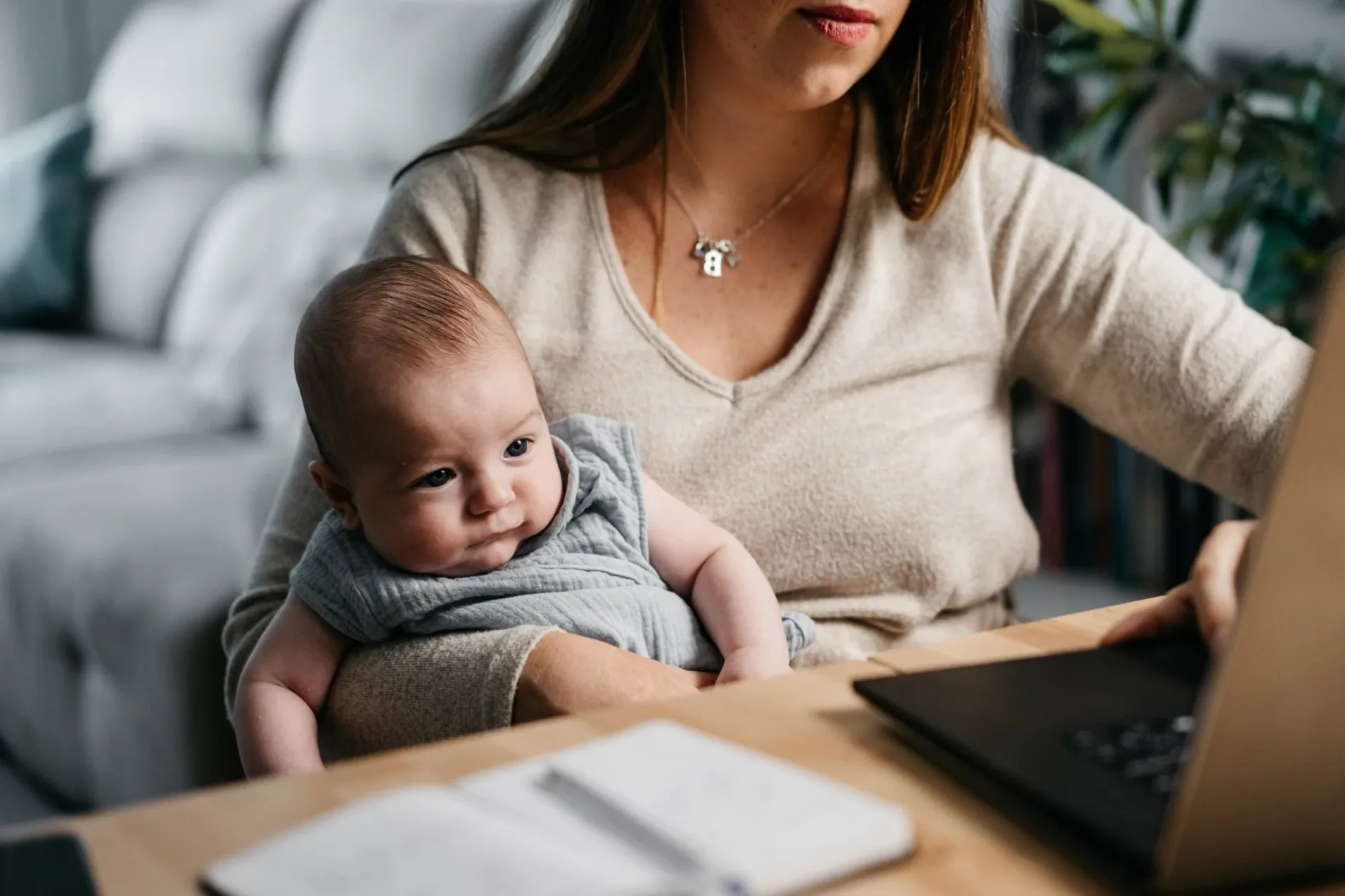 A woman working at a laptop at home, holding her baby